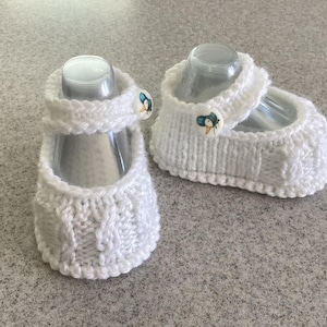 Könnte beinhalten: Ein Paar weiße gestrickte Babyschuhe mit einem weißen Knopf an jedem Schuh.