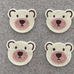 May include: Four handmade, crocheted bear face appliques. Each bear face features a white body, brown ears, eyes, and nose, and a pink muzzle. The appliques are arranged on a grey background.