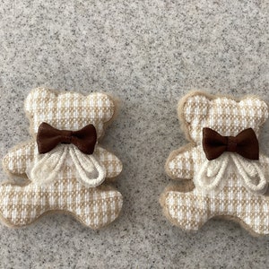 May include: Two small beige teddy bears with white checked fabric and brown bows. The bears are sitting on a light grey surface.