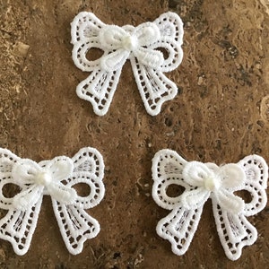 May include: Three white lace bows with a pearl centre. The bows are delicate and intricate, perfect for adding a touch of elegance to any project.