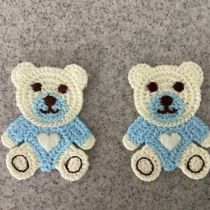 May include: Two crocheted teddy bear appliques in cream with blue and white embroidered details. Each bear has a heart on its chest.