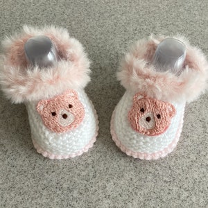 May include: A pair of white knitted baby booties with pink fur trim and a pink bear face on the front.