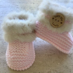 Könnte beinhalten: Ein Paar rosa gestrickte Babyschuhe mit weißem Fellbesatz. Die Schuhe haben einen Holzknopf mit dem Text "Isn't She Lovely" darauf.
