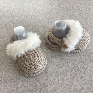 Könnte beinhalten: Ein Paar beige gestrickte Babyschuhe mit flauschigem weißem Besatz. Die Schuhe werden mit einem passenden Strickhut präsentiert, der ebenfalls mit weißem Flaum verziert ist. Das Set ist auf Wärme und Komfort ausgelegt.