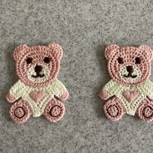 May include: Two handmade crochet teddy bear appliques. Each bear has a pink head and body with a cream-coloured shirt. The bears have brown eyes and noses, and a pink heart on the shirt. Ideal for crafting projects.