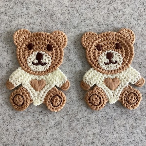 May include: Two crocheted teddy bear appliques. The bears are mainly brown with cream-coloured shirts and brown heart accents. The bears have dark brown eyes and noses. The appliques are on a grey textured surface.