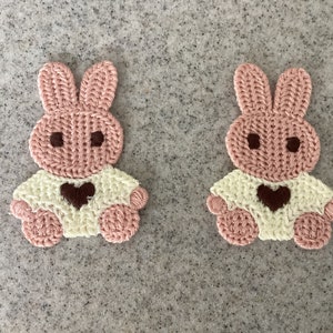 May include: Two pink crocheted bunny rabbit appliques with white crocheted shirts and brown heart accents.