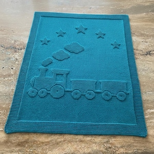 May include: A rectangular teal knitted blanket featuring a textured pattern of a steam locomotive pulling two train cars. Above the train, there are three clouds and five stars integrated into the knit design, surrounded by a wide garter stitch border.