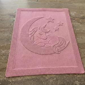 May include: A rectangular pink knitted blanket featuring a textured design of a unicorn resting on a crescent moon with three stars above it. The blanket has a wide garter stitch border surrounding the central motif of the sleeping unicorn.