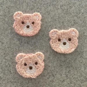 May include: Three pink bear face patches with white embroidered details and brown eyes and nose. The patches are made of a textured material and are arranged on a grey surface. These patches are ideal for sewing or crafting projects.