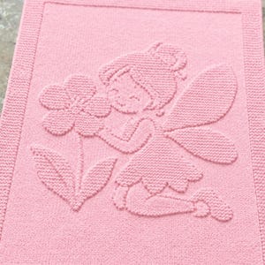 Può includere: Una coperta rettangolare in maglia rosa chiaro con un design in rilievo raffigurante una fata con ali inginocchiata accanto a un grande fiore. Il tessuto presenta un motivo a maglia testurizzato uniforme con un bordo.