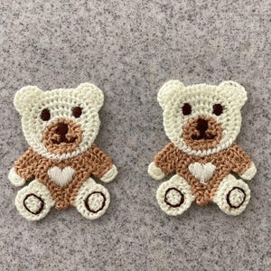 May include: Two handmade crochet teddy bear appliques. Each bear has a white head and paws, a brown body, and a white heart. The bears have black eyes and brown noses. The appliques are on a grey background.