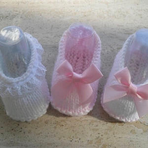 Puede incluir: Tres botines de bebé tejidos con ribete de encaje. Dos son rosas con un lazo de satén y uno es blanco con ribete de encaje.