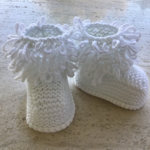 May include: A pair of white knitted baby booties with a fluffy, crocheted trim around the top.