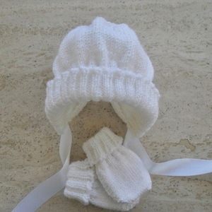 May include: White knitted baby hat and mittens set. The hat has a pointed top and a fold-over brim. The mittens are fingerless.