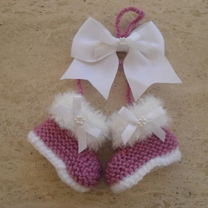 Può includere: Un paio di scarpine per neonati in maglia rosa con bordino in pelliccia bianca e fiocchi di raso bianco. Le scarpine sono appese a un fiocco di raso bianco.