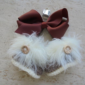May include: A pair of baby booties with a brown bow and a metal clip. The booties are white with brown and cream accents, and have wooden buttons that read "How Cute Am I".