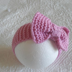 May include: A pink knitted headband with a large bow. The headband is made with a soft, textured yarn.