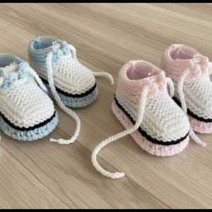 Puede incluir: Zapatillas de bebé tejidas a mano en dos pares: una en azul y blanco, la otra en rosa y blanco. Cada par presenta cordones blancos, una franja negra en la suela y un tejido de punto suave y texturizado. Estos adorables zapatos son perfectos para recién nacidos.