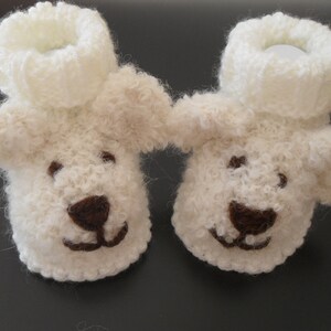 Könnte beinhalten: Ein Paar weiße gestrickte Babyschuhe mit braunen Filzgesichtern. Die Schuhe haben eine flauschige Textur und sind so gestaltet, dass sie wie kleine Hunde aussehen.