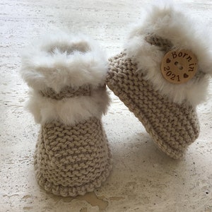 Puede incluir: Un par de botines de bebé de punto beige con ribete de piel blanca. Los botines tienen un botón de madera con el texto "Born in 2021" en ellos.