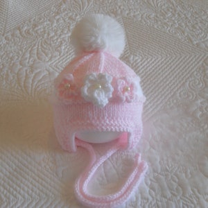 May include: A pink knitted baby hat with a white pom pom and white flower embellishments.