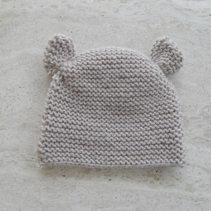 May include: A light gray knitted baby hat with two small ear flaps. The hat is made of a soft, textured yarn.