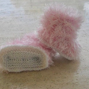 May include: A pair of pink baby booties with white knit soles. The booties are made of a fluffy, fuzzy yarn.