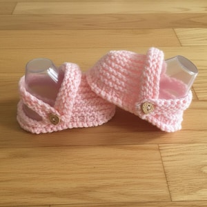 May include: A pair of light pink, hand-knitted baby sandals. The sandals have a strap that fastens with a wooden button. The sandals are displayed on a wooden surface.