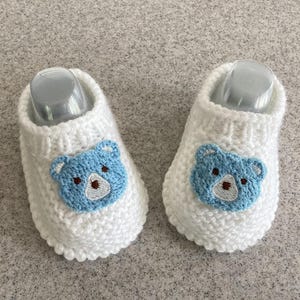 Op de afbeelding: Een paar witte gebreide babyslofjes. Elk slofje heeft een lichtblauwe berenbek met een bruine neus en geborduurde details. De slofjes zijn ontworpen om de voetjes van een baby warm te houden.