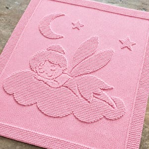 May include: A rectangular light pink knitted blanket featuring a textured, raised design of a fairy sleeping on a cloud. Above the fairy, there is a crescent moon and two small stars. The blanket has a wide, textured border around the edge.