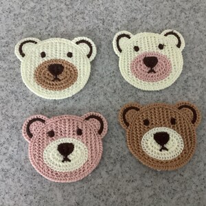 May include: Four handmade crochet bear face coasters. Two are white with brown and tan accents, one is pink with white and brown accents, and the last is brown with white and tan accents. The coasters are arranged on a grey surface.