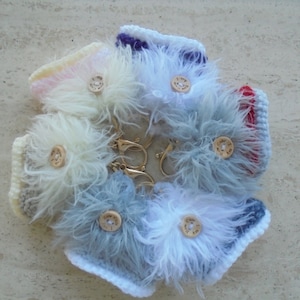 May include: Seven fluffy keychains with different colored crocheted bases and white fur. Each keychain has a gold button with a different inscription.