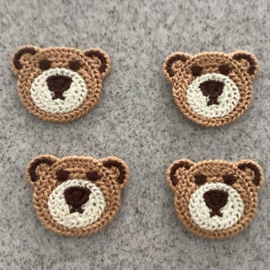May include: Four crocheted bear face appliques. Each bear face is brown with a white muzzle and dark brown details for the eyes, nose, and ears. The appliques are arranged on a grey surface.
