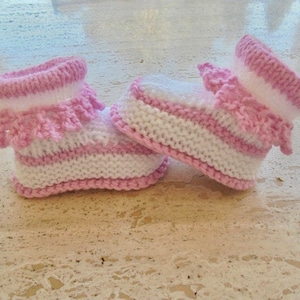 May include: A pair of pink and white knitted baby booties with a ruffled trim.