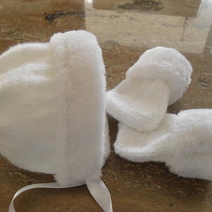 May include: White knitted baby hat and booties with a fluffy white trim. The hat has a white ribbon tied under the chin.