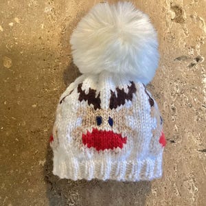 Op de afbeelding: Een witte gebreide muts met een rendiergezichtsontwerp. De muts heeft een pluizige witte pompon bovenop, bruine geweien, een bruin gezicht, blauwe ogen en een rode mond. Deze wintermuts is ideaal voor de feestdagen.