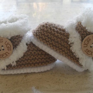 May include: A pair of baby booties with a brown knitted exterior and white faux fur trim. Each bootie features a wooden button with the "UGG" logo. The booties are designed to keep a baby's feet warm and cosy.