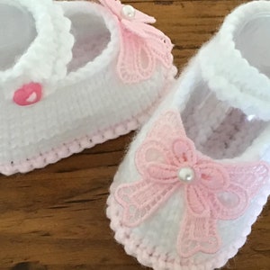 Puede incluir: Un par de zapatos de bebé de ganchillo blanco con detalles en rosa. Los zapatos presentan un estilo Mary Jane con una correa y un delicado lazo de encaje rosa con un centro de perlas. Los bordes están ribeteados en rosa.