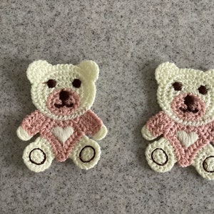 May include: Two crocheted teddy bear appliques made with pink and white yarn. The bears have brown embroidered eyes and noses and a white heart on their chests.