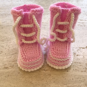 May include: A pair of pink knitted baby booties with white laces. The booties have a white trim around the bottom.