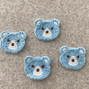 May include: Four light blue, embroidered bear patches. Each patch features a white muzzle, brown nose, and white ear details. The patches are arranged on a textured, grey surface.