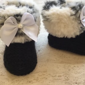 May include: A pair of black baby booties with white faux fur trim and white satin bows. Each bow has a small pearl accent.