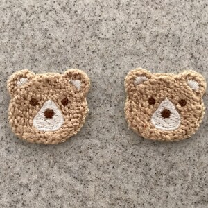 May include: Two embroidered bear patches. Each patch features a tan bear face with a white muzzle, brown nose, and small brown eyes. The patches are made of a textured, woven material and are set against a grey background.