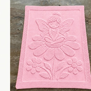 May include: A rectangular light pink knitted blanket featuring a raised texture design of a fairy sitting on a large flower, flanked by two smaller flowers with leaves. The border consists of a simple garter stitch pattern framing the central motif.