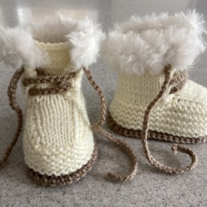 Könnte beinhalten: Ein Paar cremefarbene, gestrickte Babyschuhe mit braunen Verzierungen und Schnürsenkeln. Die Oberseite jedes Schuhs ist mit flauschigem, weißem Kunstfell verziert. Die Schuhe sind so konzipiert, dass sie die Füße eines Babys warm halten.