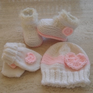 May include: A set of knitted baby clothes in white and pink. The set includes a hat, booties, and mittens. Each piece has a pink heart-shaped embellishment.