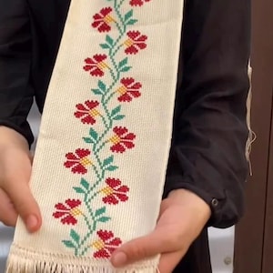 May include: Cream-colored embroidered scarf featuring a floral pattern with red flowers, green leaves, and yellow centers. The scarf has a fringed edge. The design is centered on the scarf.