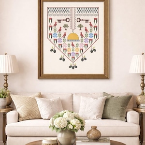 May include: A framed cross-stitch artwork featuring a heart-shaped design with keys, birds, and floral elements in red, blue, yellow, and green. The artwork is displayed above a white sofa with decorative pillows and a wooden coffee table.