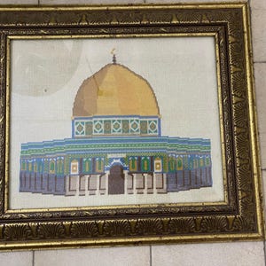 May include: Cross-stitch depiction of the Dome of the Rock in Jerusalem, presented in a gold-coloured, ornate wooden frame. The artwork showcases a gold dome, blue and green details, and intricate architectural patterns. The frame provides a traditional aesthetic.